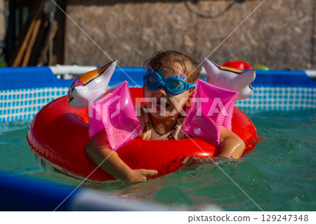 Happy little girl swimming and playing in a home pool, wearing inflatable armbands and a bright red swim ring, enjoying carefree moments during summer holidays under the sunny sky. Happy little girl swimming and playing in a home pool, wearing inflatable armbands and a bright red swim ring, enjoying carefree moments during summer holidays under the sunny sky. 129247348