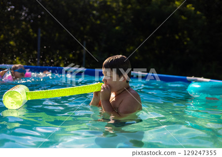 A boy blows up a toy while swimming next to a girl in a colorful backyard pool, enjoying summer days filled with laughter, splashes and playful moments. Children swimming in the pool. A boy blows up a toy while swimming next to a girl in a colorful backyard pool, enjoying summer days filled with laughter, splashes and playful moments. Children swimming in the pool. 129247355