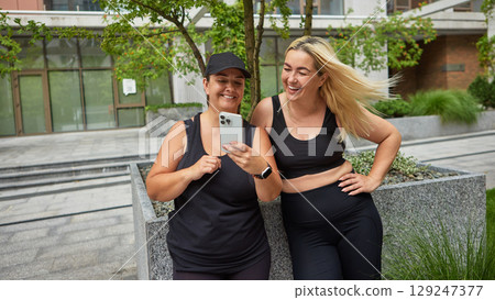 Two women in black sportswear watching video on phone while resting 129247377