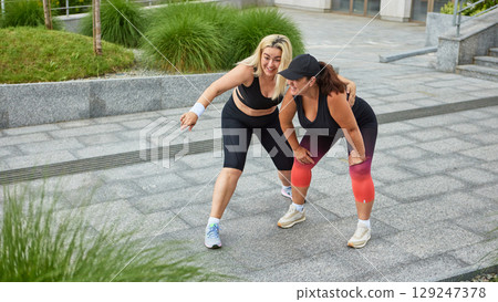 Two sporty women laughing while bending over during outdoor workout 129247378