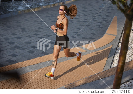 Female running on city walkway with sports attire and sunglasses during morning hours Female running on city walkway with sports attire and sunglasses during morning hours 129247418
