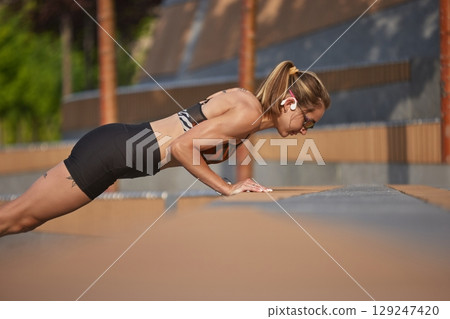 Woman executing push-ups outside on a bright day with concentration and resolve 129247420
