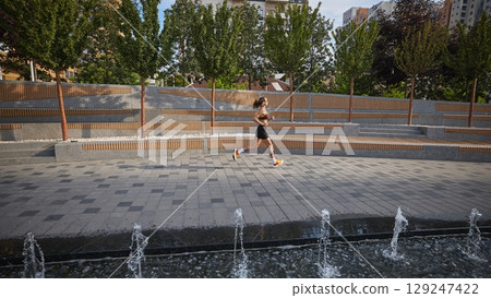 Woman jogging in urban park with fountains and greenery on sunny day Woman jogging in urban park with fountains and greenery on sunny day 129247422