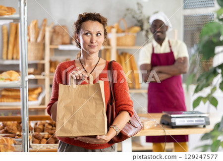Woman buyer with pastry and baguettes in paper package stands near glass showcase in bakery 129247577