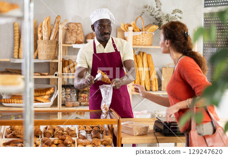 African man entrepreneur serves female customer, puts croissant in disposable plastic bag 129247620