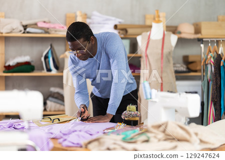 Middle-aged tailor cutting fabric with scissors on worktable Middle-aged tailor cutting fabric with scissors on worktable 129247624