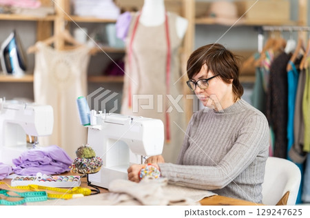 Working in sewing workshop - female dressmaker working at sewing machine making stitches on fabric Working in sewing workshop - female dressmaker working at sewing machine making stitches on fabric 129247625