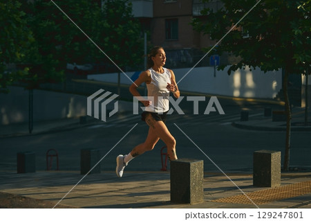 Female jogger in the early morning running along an urban street wearing casual sportswear Female jogger in the early morning running along an urban street wearing casual sportswear 129247801