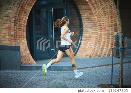 Woman with tattoos running past curved brick wall in white top and black shorts Woman with tattoos running past curved brick wall in white top and black shorts 129247805