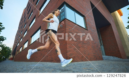 Tattooed female athlete running near red brick building in white tank top and black shorts 129247826