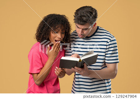 Interracial couple reading book with surprised expressions on beige background Interracial couple reading book with surprised expressions on beige background 129247904