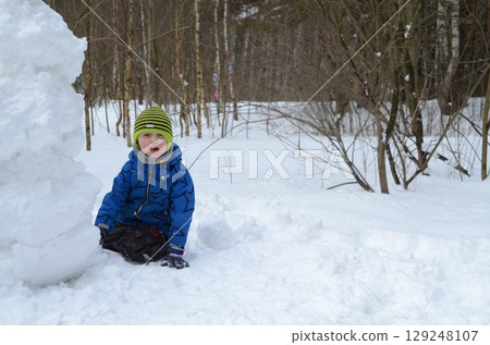 Child Playing in Snow Near Large Snowman in Winter Forest 129248107