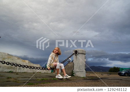 Child Relaxing on Urban Chain Fence with Overcast Sky 129248112