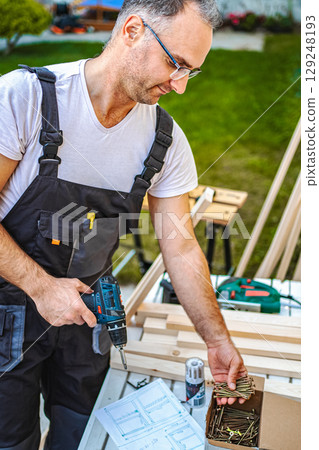 Mature male carpenter using electric screwdriver on a wooden plank outdoors 129248193