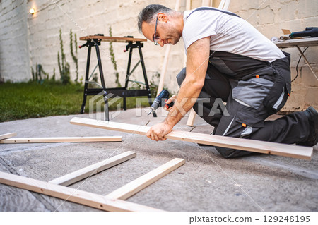 Mature male carpenter using electric screwdriver on a wooden plank outdoors 129248195