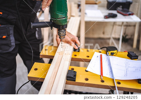 Unrecognizable male carpenter using electric drill on a wooden plank outdoors 129248205