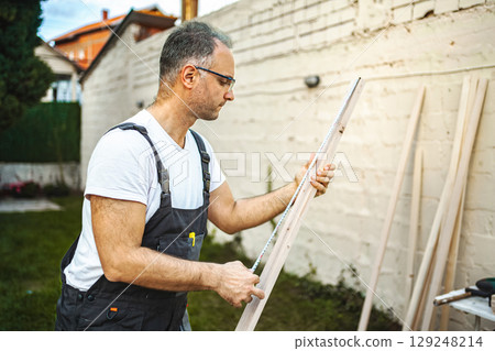 Mature male carpenter measuring wooden plank outdoors Mature male carpenter measuring wooden plank outdoors 129248214