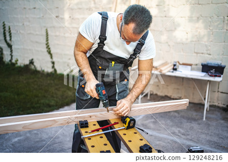 Mature male carpenter using electric drill on a wooden plank outdoors 129248216