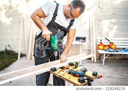 Mature male carpenter using electric drill on a wooden plank outdoors 129248219