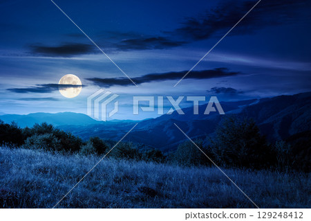 mountain landscape with forest on the hill in autumn at night. beautiful view in to the rural valley in full moon light. alpine countryside scenery under sky with clouds 129248412