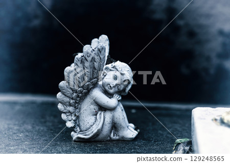 Vintage dirty statue angel on tomb stone in old cemetery. Small winged angelic figurine on dark grave yard Vintage dirty statue angel on tomb stone in old cemetery. Small winged angelic figurine on dark grave yard 129248565