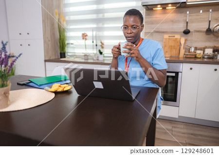 Nurse drinking coffee and working from home using laptop computer 129248601