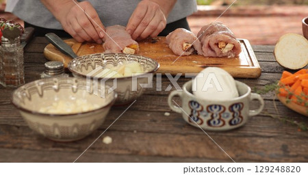 Turkey roll preparation process featuring a chef carefully stuffing turkey cuts with flavorful ingredients on a wooden countertop surrounded by fresh vegetables and seasonings 129248820