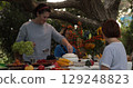 Celebration of Thanksgiving dinner with family gathering around a beautifully arranged table filled with seasonal dishes and vibrant decorations under a large tree outdoors 129248823