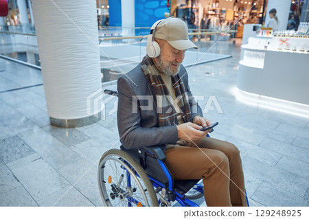 Elderly man comfortably seated in a wheelchair using his smartphone in a modern mall setting 129248925