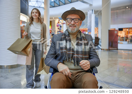 Mature man in a wheelchair is assisted by a woman in a shopping mall 129248930