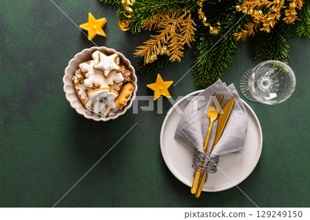 A festive table setting with an empty plate, napkin and cutlery and gingerbread on a green background. Spruse branches and golden tree ornaments enhance the holiday ambiance, creating a cozy vibe A festive table setting with an empty plate, napkin and cutlery and gingerbread on a green background. Spruse branches and golden tree ornaments enhance the holiday ambiance, creating a cozy vibe 129249150