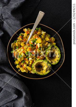 A colorful salad combines boiled egg, tender chicken breast, diced potatoes, vibrant carrots, and fresh green peas, topped with creamy avocado and sesame seeds, all drizzled with olive oil 129249211