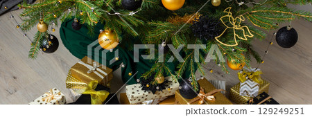 Sophisticated gold and black-toned gift boxes paired with glowing candles and intricate ornaments, nestled under the Christmas tree for a warm and modern holiday celebration, banner 129249251