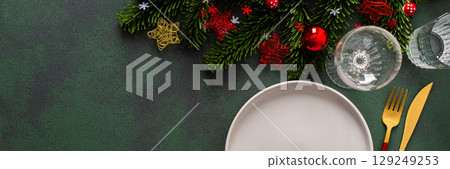 A cozy Christmas table with an empty plate and cutlery on a rich green background. Red Christmas tree decorations and spruse branches complete the festive look, banner A cozy Christmas table with an empty plate and cutlery on a rich green background. Red Christmas tree decorations and spruse branches complete the festive look, banner 129249253