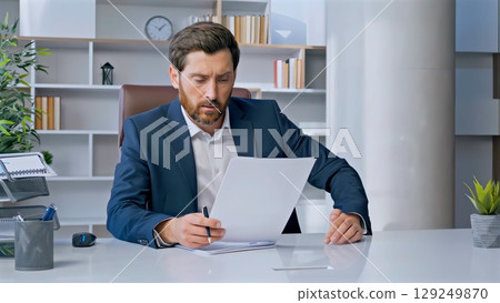 Businessman dressed in a tailored suit and tie, sitting at a modern office desk, intently reading documents while holding a pen, deeply focused on the important paperwork 129249870