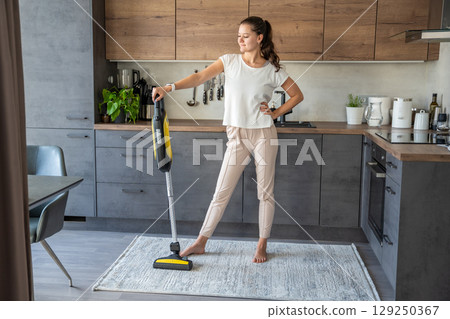 Young woman after vacuuming carpet with wireless vacuum cleaner in kitchen. Household and useful technology concept. Young woman after vacuuming carpet with wireless vacuum cleaner in kitchen. Household and useful technology concept. 129250367