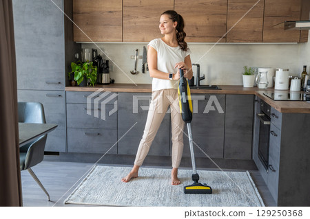 Woman vacuuming carpet with wireless vacuum cleaner in kitchen. Household and useful technology concept.  129250368