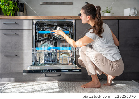Young woman removes clean ceramic dishes from dishwasher. Household and useful technology concept. Young woman removes clean ceramic dishes from dishwasher. Household and useful technology concept. 129250370