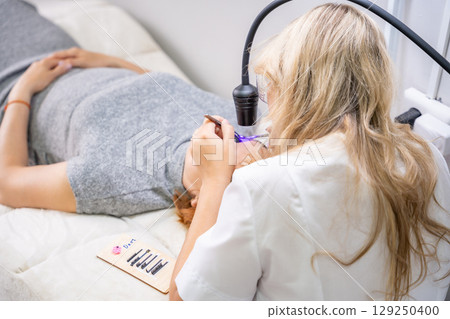 Eyelash extension procedure using ultraviolet lamp in modern beauty salon. Innovative lash enhancement technology with precise application technique. Eyelash extension procedure using ultraviolet lamp in modern beauty salon. Innovative lash enhancement technology with precise application technique. 129250400