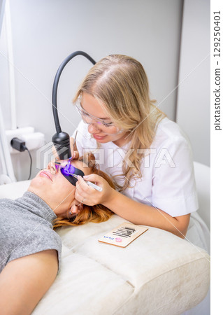 Eyelash extension procedure using ultraviolet lamp in modern beauty salon. Innovative lash enhancement technology with precise application technique. 129250401