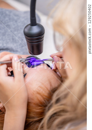 Close up view of eyelash extension procedure using ultraviolet lamp in modern beauty salon. Innovative lash enhancement technology with precise application technique. 129250402