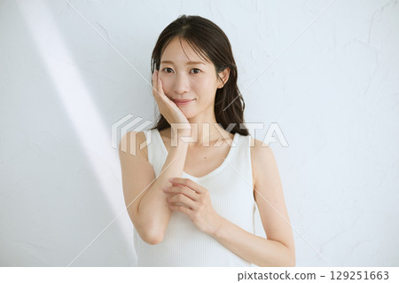 Smiling young woman touching her skin and doing skin care - Natural light, white background 129251663