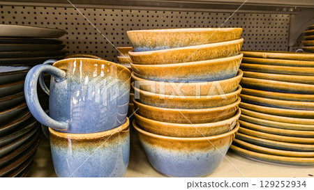 Handcrafted ceramic bowls and mugs with blue and brown glaze, stacked on store shelf. Suitable for concepts of artisan pottery, home goods and kitchenware Handcrafted ceramic bowls and mugs with blue and brown glaze, stacked on store shelf. Suitable for concepts of artisan pottery, home goods and kitchenware 129252934