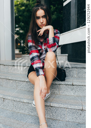 Woman in a plaid shirt and black shorts sits on a set of stairs Woman in a plaid shirt and black shorts sits on a set of stairs 129252944