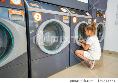 Daily life moment at a self-service laundry, child observing the washing process while waiting. Accessibility of public laundry services and everyday family routine. 129253773