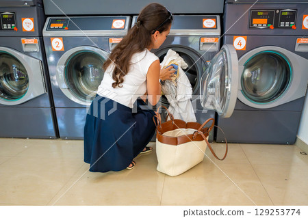 Young woman loading laundry into washing machine in self-service laundry facility. Everyday routine of washing clothes in modern public laundromat. Young woman loading laundry into washing machine in self-service laundry facility. Everyday routine of washing clothes in modern public laundromat. 129253774