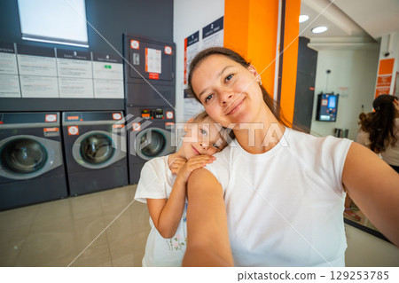 Mother and daughter taking a selfie while waiting in a self-service laundromat, looking at camera and bonding during laundry time, promoting shared responsibilities and everyday routine Mother and daughter taking a selfie while waiting in a self-service laundromat, looking at camera and bonding during laundry time, promoting shared responsibilities and everyday routine 129253785