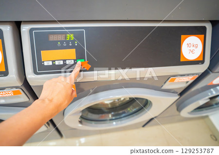 Close-up of female hand selecting washing program on modern washing machine. Choosing laundry mode in public self-service facility. Close-up of female hand selecting washing program on modern washing machine. Choosing laundry mode in public self-service facility. 129253787