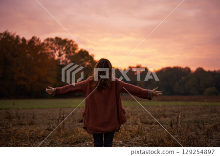 In a tranquil autumn setting, a woman stands in a field, arms outstretched, embracing the vibrant colors of the sunset and the warmth of the season as trees glow in the background 129254897