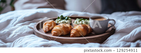 A wooden tray holds three buttery croissants and two small bowls of green dip, accompanied by a steaming cup of coffee, resting on soft bedding, creating a tranquil breakfast setting 129254911